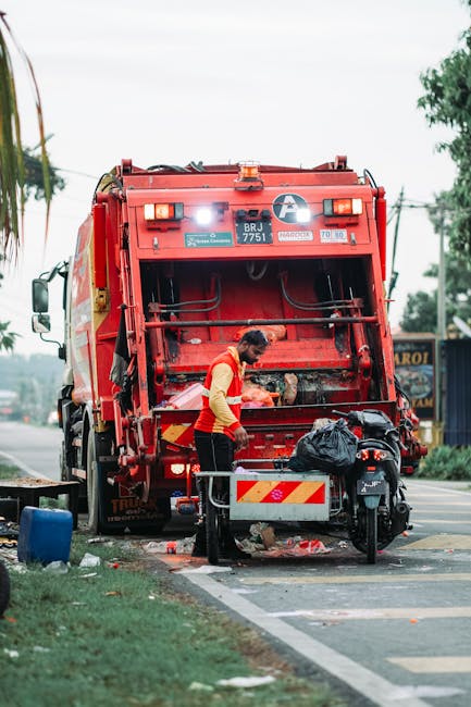 A waste collection worker is standing on a paved roadside area in front of a large red rubbish truck, which is aligned with a curb and partially blocking the temporary parking lane. The worker, dressed in a high-visibility yellow and red vest, is monitoring or operating the rear-loading mechanism of the truck, which is open to reveal internal components used for waste collection. A large black plastic bag, filled with rubbish, is placed on the ground near the worker, while additional small debris and waste materials are scattered on the pavement. Nearby, a black motorbike with a small black box attached to the rear is parked, and there is a metal barrier with an orange and white striped reflective panel situated close to the worker. The background features blurred images of trees, traffic lights, utility poles, and signs, indicating an urban or suburban setting. The scene depicts an independent waste collection operation, typical of private rubbish removal services, with natural daylight illuminating the area and emphasizing the textures of the waste materials and equipment.