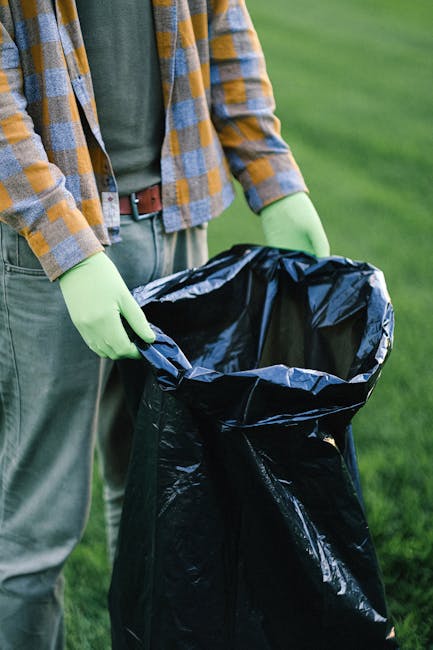 A person, partially visible from the shoulders down, is wearing a grey-green shirt, a plaid jacket in shades of yellow, grey, and blue, and grey trousers secured with a brown belt. They are standing outdoors on a grassy surface, holding open a large black trash bag lined with a silver interior. The individual’s hands are covered with light green disposable gloves, and they are in the process of collecting or disposing of waste. The background features a blurry expanse of vibrant green grass or lawn, suggesting a park, garden, or outdoor area. The scene emphasizes proper waste handling in an outdoor environment, consistent with rubbish removal activities, with no additional objects or vehicles visible in the frame. The lighting is natural, with soft shadows indicating daylight, and the overall composition reflects routine on-site waste collection or cleanup activities, aligning with private or council-led rubbish management efforts.