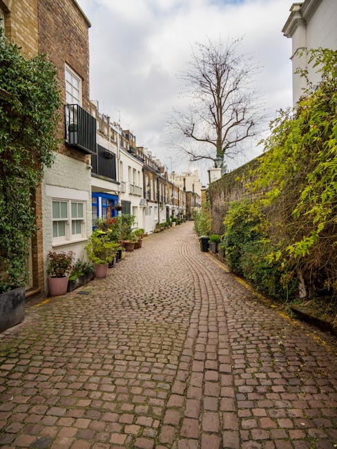 A quiet residential street in West Kensington lined with parked cars on both sides, including compact hatchbacks, sedans, and small SUVs, all neatly positioned within designated parking bays. The left side of the street features a row of terraced Victorian-style houses with white facades, sash windows, and decorative cornices, while the right side shows similar residential buildings with slight variations in design. Prominent in the scene are several mature cherry blossom trees with dark, gnarled branches extending horizontally across the street, heavily laden with clusters of light pink flowers that create a soft, colorful canopy overhead. Some petals have fallen onto the asphalt, scattered near the curb edges. The overcast sky is partly cloudy with patches of blue visible, casting diffuse natural light that highlights the vibrant blossoms and the textures of the vehicles and building facades. This scene subtly illustrates typical urban street parking conditions and the seasonal bloom of flowering trees, which can be associated with private or alternative waste handling scenarios in residential areas, such as on-site clearance or independent rubbish removal services, as offered by local providers like rubbishremovalwestkensington.org.uk.