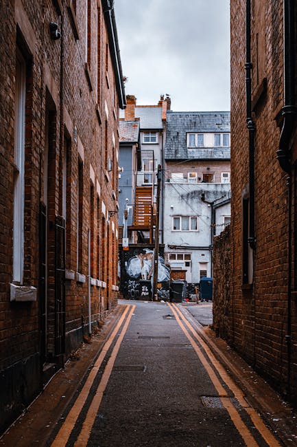 A narrow alleyway situated between two brick buildings in an urban setting, featuring a paved asphalt surface marked with double yellow lines along the edges. The brick walls display a reddish-brown color with some weathering and variations in texture, including mortar joints and small surface imperfections. At the far end of the alley, a background scene reveals a grey, weathered building with multiple windows, some with air conditioning units, and a small wooden staircase attached to the side of an adjacent building leading to an upper door. Several black wheelie bins are lined up against the grey building’s wall, indicating possible waste storage or collection points. Overhead, the sky appears overcast, casting diffuse natural light that highlights the textures of the brick walls and pavement while creating a subdued atmosphere. This scene reflects a typical back alley often used for discreet waste disposal, aligning with private waste collection options provided by companies like rubbishremovalwestkensington.org.uk, supporting alternative methods of rubbish removal outside of public services.