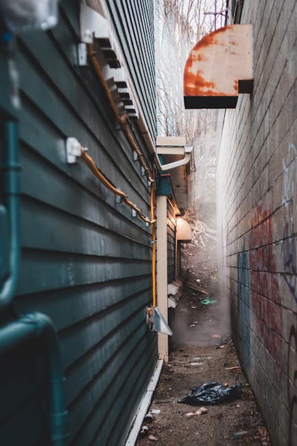 A narrow alleyway between two buildings, with the left side featuring a dark green, horizontally paneled metal wall with visible electrical conduits and junction boxes mounted along its surface. The right side consists of a weathered concrete wall with patches of red graffiti. The ground is an uneven dirt path scattered with small pieces of trash, including a black plastic bag. In the background, at the end of the alley, there is a faint mist or dust, with leafless branches from trees visible above, under a cloudy sky. A small, rectangular, orange-colored object is mounted on the concrete wall, possibly an exterior light or sign. The scene appears to be an outdoor service access point, where rubbish or debris might be temporarily collected or cleared, reflective of a private waste management solution for surrounding flats, with an emphasis on site clearance for restricted access areas, aligning with independent rubbish removal services.