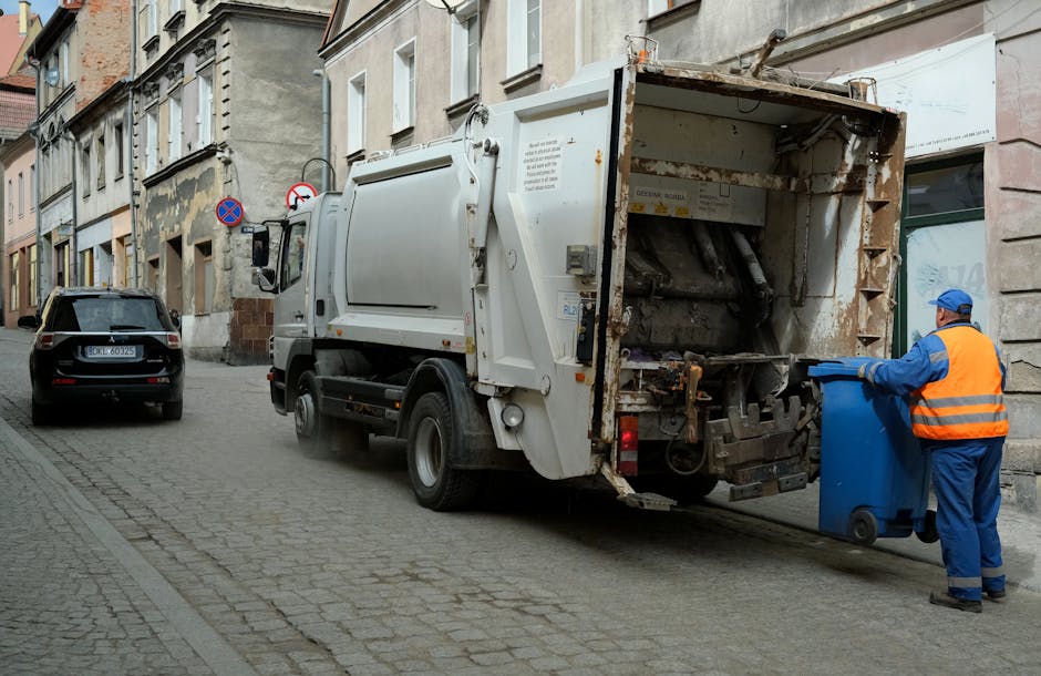 A street scene depicts a waste collection vehicle positioned adjacent to a sidewalk in an urban area. The vehicle is a large, white, industrial garbage truck with a rusted rear compactor section, which is partially open, revealing mechanical components and a textured metal surface with signs of wear. A worker dressed in blue protective clothing, including a high-visibility orange vest and matching headgear, is standing to the right of the truck, holding a blue wheeled waste bin and preparing to unload or empty it into the vehicle. Behind the truck, a dark grey car with a European license plate is parked parallel to the curb, and further along the street, there are multistory buildings with beige and grey stone facades, some featuring small balconies and large windows. The street is paved with grey cobblestones, and a no parking sign is visible mounted on a pole on the building frontage. The environment appears to be a typical city setting for private waste collection, with surrounding urban architecture and street furniture supporting the context of rubbish removal services by companies like rubbishremovalwestkensington.org.uk.