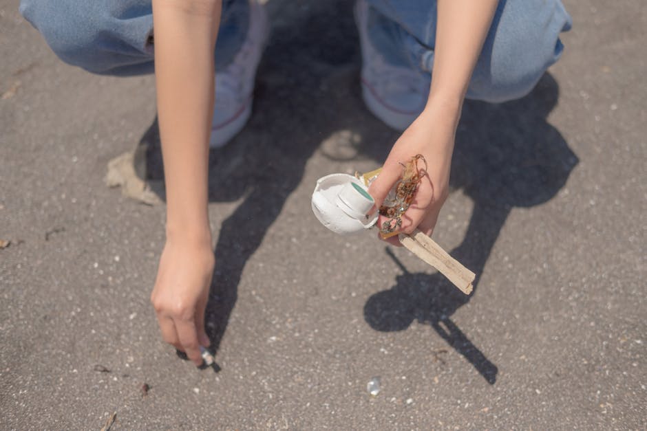 A person wearing light blue jeans and white sneakers is crouching on a paved surface outdoors, holding a small broken object assembled from various discarded materials. The object includes a white foam piece, possibly an egg carton segment, attached to a small wooden stick or dowel. The person's right hand gently grasp the object, while the left hand is positioned near the ground, with fingers slightly curled. The background shows a textured, grey asphalt surface with small stones and debris scattered around. The scene appears to be part of an outdoor activity or cleanup related to waste removal or environmental awareness, reflecting themes of waste collection and proper disposal. The lighting is natural, illuminating the scene evenly, with shadows cast by the person and the object they are holding, emphasizing the textures of the materials and the paved environment.