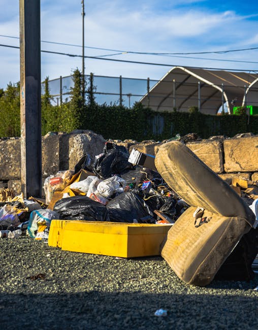 A street scene depicts a waste collection vehicle positioned adjacent to a sidewalk in an urban area. The vehicle is a large, white, industrial garbage truck with a rusted rear compactor section, which is partially open, revealing mechanical components and a textured metal surface with signs of wear. A worker dressed in blue protective clothing, including a high-visibility orange vest and matching headgear, is standing to the right of the truck, holding a blue wheeled waste bin and preparing to unload or empty it into the vehicle. Behind the truck, a dark grey car with a European license plate is parked parallel to the curb, and further along the street, there are multistory buildings with beige and grey stone facades, some featuring small balconies and large windows. The street is paved with grey cobblestones, and a no parking sign is visible mounted on a pole on the building frontage. The environment appears to be a typical city setting for private waste collection, with surrounding urban architecture and street furniture supporting the context of rubbish removal services by companies like rubbishremovalwestkensington.org.uk.
