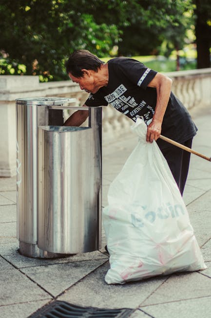 A middle-aged woman with short dark hair, wearing a black t-shirt with white and blue text and black trousers, is seen disposing of rubbish into a cylindrical stainless steel litter bin. She leans slightly forward, holding a large white trash bag filled with waste in her left hand, while her right hand is extended into the bin to deposit the rubbish. The bin is situated on a paved sidewalk, bordered by a low stone wall with a decorative balustrade. In the background, lush green trees and foliage provide a natural setting, contrasting with the urban environment. The scene is illuminated by natural daylight, highlighting the reflective surface of the stainless steel bin and the textured pavement beneath her feet. The woman's actions depict a scene of proper waste disposal, relevant to independent rubbish removal or on-site clearance, supported by services such as those offered by rubbishremovalwestkensington.org.uk, emphasizing the importance of responsible waste management in local outdoor environments.