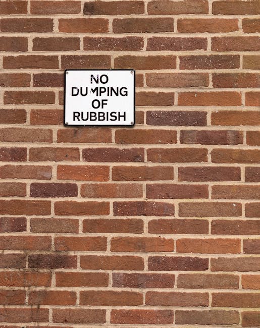 A vertically oriented rectangular metal sign mounted on a red brick wall, with a white background and black lettering that reads 'NO DUMPING OF RUBBISH'. The sign appears weathered, with some minor scratches and faded areas around the edges. The brick wall behind the sign is composed of evenly spaced bricks in various shades of reddish-brown, with light mortar joints creating a uniform pattern. The lighting is natural, casting soft shadows below the sign and against the brick surface. The scene suggests an environment where private waste removal or on-site clearance is regulated, with the sign serving as a clear directive against illegal dumping, which is relevant to rubbish removal services such as those provided by companies operating in West Kensington and North End Road areas.
