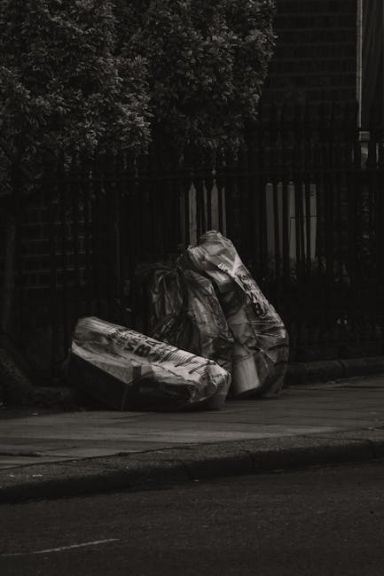 A collection of black and white plastic bags and large trash bags filled with waste, placed on the edge of a pavement in front of a black metal fence with vertical bars. The bags appear to contain various discarded materials and are loosely tied, with some showing visible creases and folds. Behind the fence, a dense cluster of leafy trees partially obscures the background, which appears to be a building or structure covered in a dark material, possibly wood or metal. The scene is illuminated by natural light, creating subtle shadows on the bags and pavement, highlighting the textures of the plastic and the concrete surface. The overall setting suggests an outdoor area designated for rubbish disposal, possibly as part of a private collection or external waste removal service, supporting the concept of independent waste handling and rubbish removal in West Kensington.