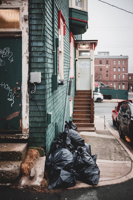 A row of black rubbish disposal bags, filled and tied, are stacked near the entrance of a green wooden building with red and white trim, situated on an urban street. The bags rest on a concrete pavement adjacent to a small section of rough, partially melting snow and a worn concrete step leading to a stairway with wooden steps and brown railings, which ascends to a white door with a decorative overhang above. To the left, part of an aged, graffiti-covered door or wall is visible, showing signs of wear and peeling paint. In the background, parked cars, including a black vehicle, are aligned along the street, with a brick warehouse or industrial building further back under a lightly overcast sky. Exterior electrical boxes and wires are mounted on the building’s façade, indicating typical urban infrastructure. The scene reflects typical private waste management practices away from municipal collection, aligning with independent rubbish removal efforts in West Kensington, as managed by services such as those offered by rubbishremovalwestkensington.org.uk.