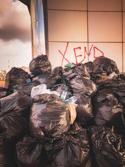 A row of black rubbish disposal bags, filled and tied, are stacked near the entrance of a green wooden building with red and white trim, situated on an urban street. The bags rest on a concrete pavement adjacent to a small section of rough, partially melting snow and a worn concrete step leading to a stairway with wooden steps and brown railings, which ascends to a white door with a decorative overhang above. To the left, part of an aged, graffiti-covered door or wall is visible, showing signs of wear and peeling paint. In the background, parked cars, including a black vehicle, are aligned along the street, with a brick warehouse or industrial building further back under a lightly overcast sky. Exterior electrical boxes and wires are mounted on the building’s façade, indicating typical urban infrastructure. The scene reflects typical private waste management practices away from municipal collection, aligning with independent rubbish removal efforts in West Kensington, as managed by services such as those offered by rubbishremovalwestkensington.org.uk.