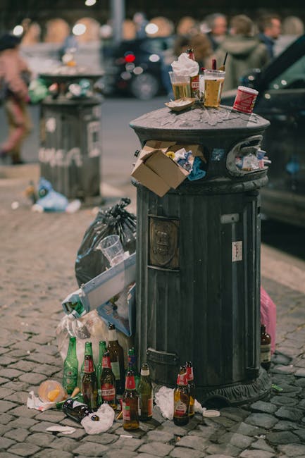 A black, cylindrical publicly accessible rubbish bin with a domed lid, situated on a cobblestone pavement in an urban environment, showing signs of overflow and litter accumulation. The top of the bin is cluttered with discarded food containers, cups with plastic lids, and a crumpled napkin, some of which are resting on the lid's surface. Around the base, there are numerous glass bottles, including beer bottles in green and brown, with some lying on their sides and a few amid crumpled paper and plastic rubbish, indicating improper disposal. In the background, blurred pedestrians, parked cars, and a second rubbish bin are visible, adding to the busy street scene. The environment appears to be in daylight, with shadows cast by nearby objects, and the overall scene reflects typical urban waste issues, highlighting the importance of effective rubbish removal services such as those offered by companies managing waste in West Kensington.