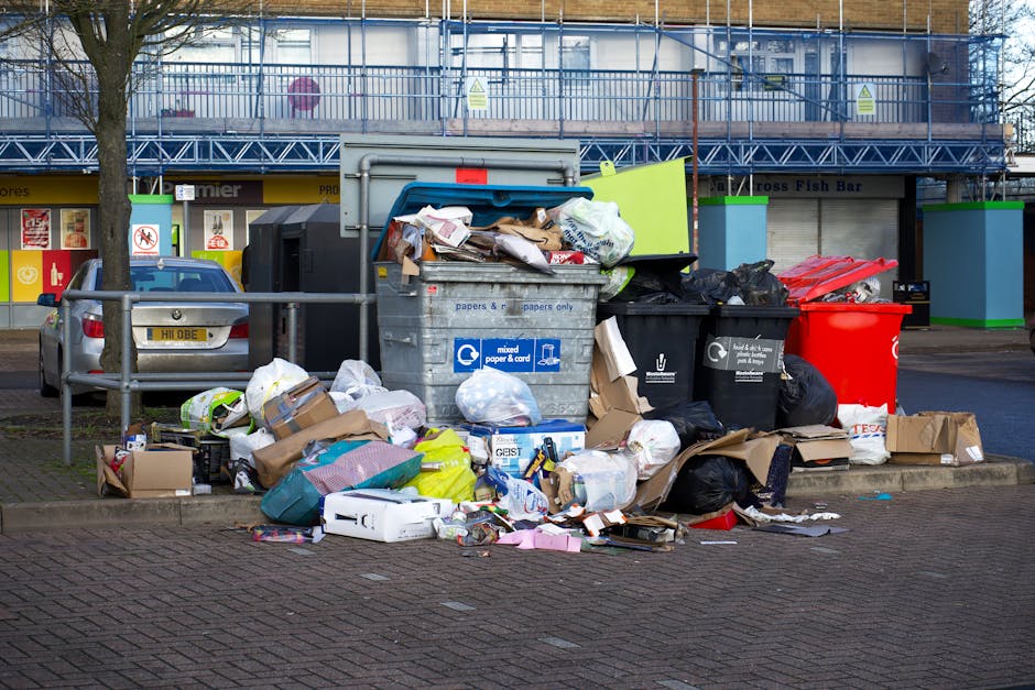 A large pile of mixed household waste and recyclables is accumulated on a paved street corner, situated in front of a commercial building with a blue scaffolding structure and signage in the background. The waste collection includes cardboard boxes, plastic wrapping, paper bags, black and green refuse bins, and loose trash spilling onto the pavement. The cardboard boxes vary in size and are flattened or partially assembled, with some torn or crumpled. The black and green bins appear to be designated for different types of waste, with labels indicating their intended contents. Additionally, there is a large, open grey recycling bin filled with papers and cardboard, positioned amidst the other rubbish, suggesting an illegal or unmanaged rubbish accumulation typical of private waste collection scenarios. To the left, a parked silver car is partially visible behind a metal guardrail, and roadside trees provide some natural elements to the otherwise cluttered scene. The overall lighting is natural, indicating daytime, emphasizing the disorder and volume of waste arranged temporarily on the street, ready for collection or disposal services, such as those provided by rubbish removal specialized companies in West Kensington.